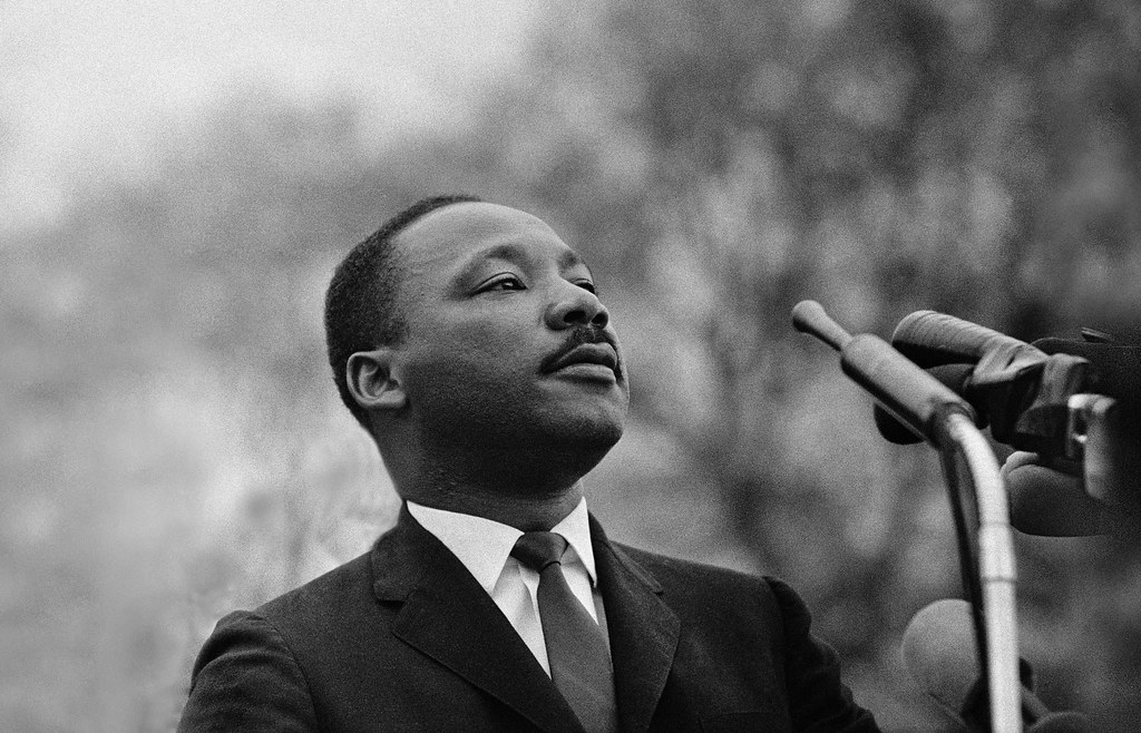 Black and white picture of MLK Jr. in a suit and tie in front of microphones