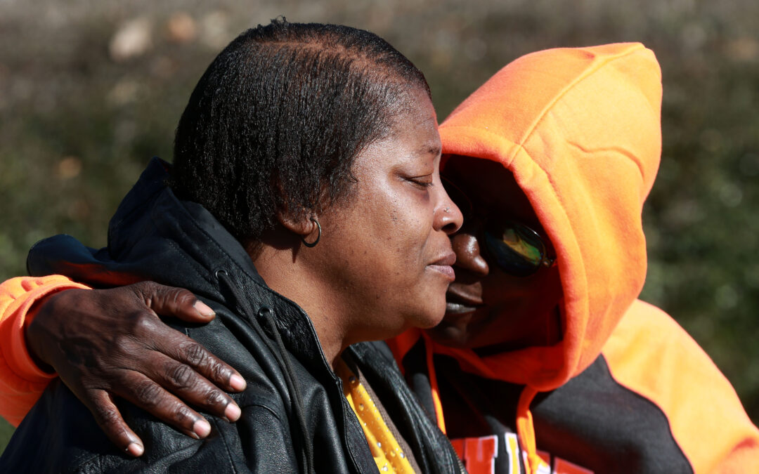 Darryl William's mother, a Black woman wearing a black jacket, closes her eyes as another Black person in an orange hoody and sunglasses puts a comforting arm around her shoulder.