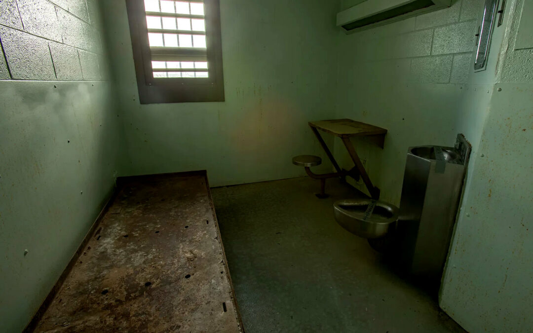 A cramped solitary confinement cell with green-tinged cinderblock walls, a barred window, rusted metal bed, small desk, toilet attached to sink, mirror, and HVAC unit.