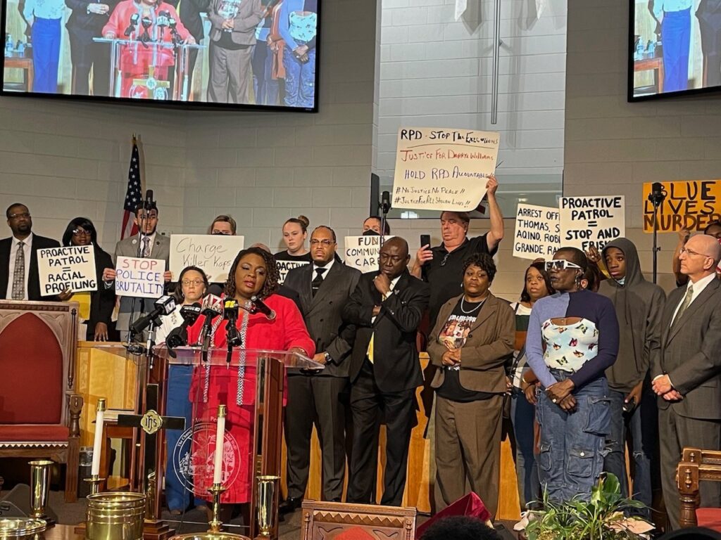 Dawn, a Black woman wearing a red dress, stands at a podium speaking in front of a group of people onstage holding signs reading "Stop Police Brutality" and "Proactive Patrol = Racial Profiling" and more.