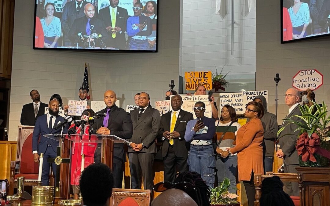 Kerwin, a Black man with shaved head and in a black suit with purple tie, stands at a podium in front of a group of people onstage holding signs reading "Blue Lives Murder" and "Stop Proactive Policing" and before an audience, of which a few heads appear in the foreground of the image.