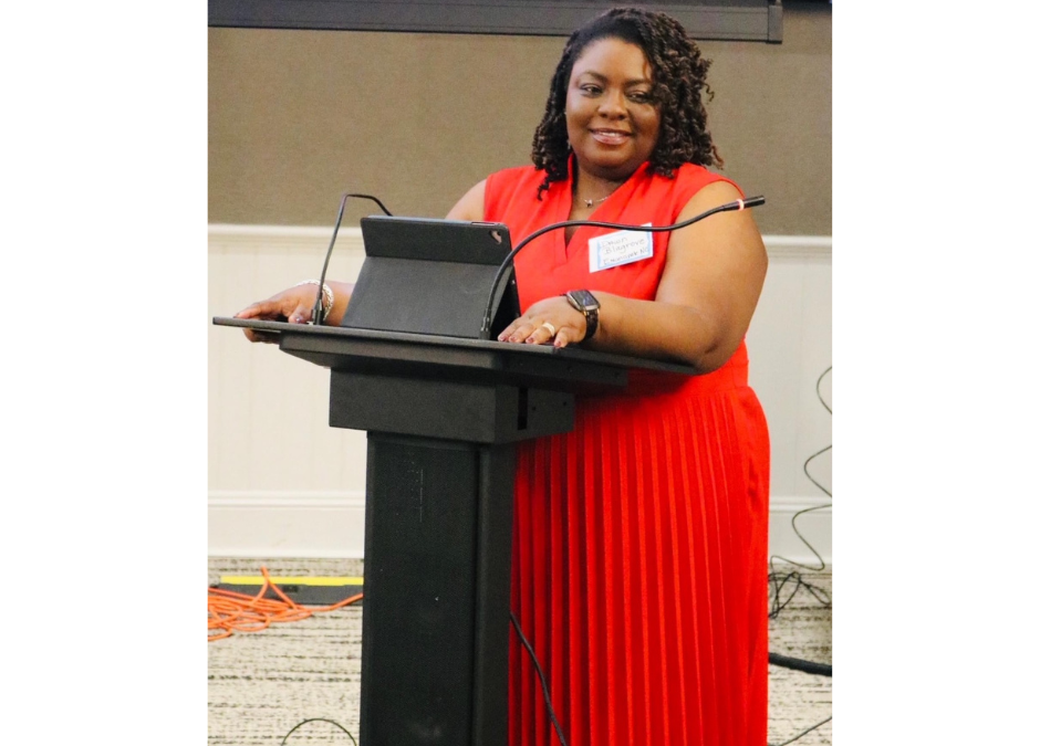 Dawn Blagrove, a Black woman in a red dress, stands smiling at a podium at the NC CRED symposium